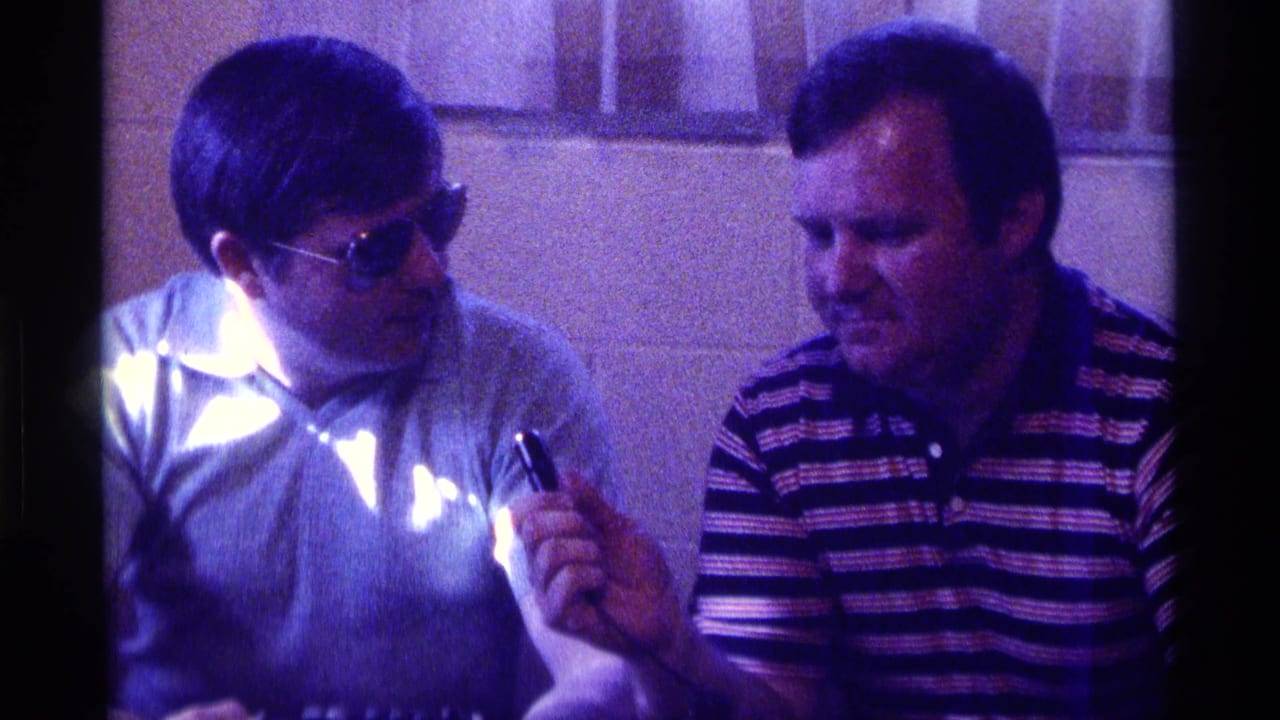 Man in sunglasses looks toward a man in a striped polo while holding a small object between them.