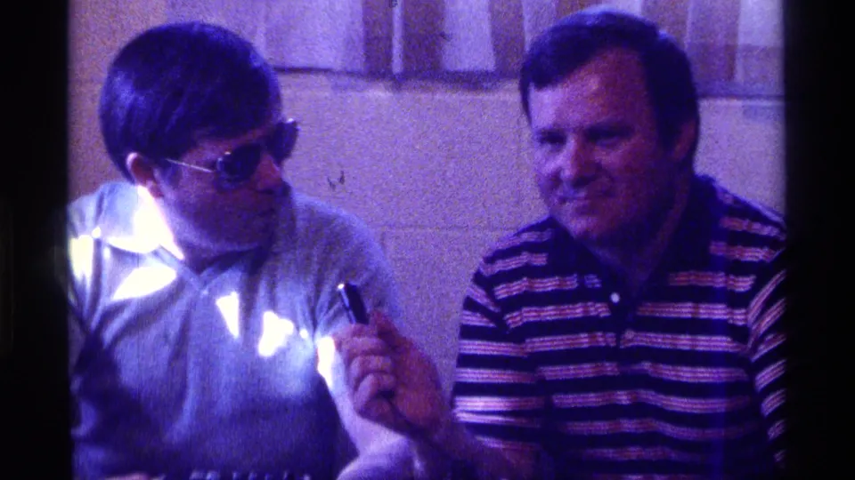 1976 Kentucky 8mm frame of two adult men seated indoors, one glancing aside while the other faces forward in a striped polo.