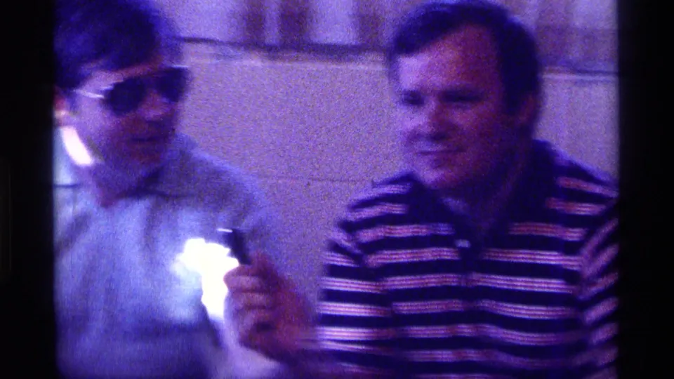 Soft, blurred 1976 Kentucky 8mm frame with the man in sunglasses moving as the striped-shirted man remains seated indoors.