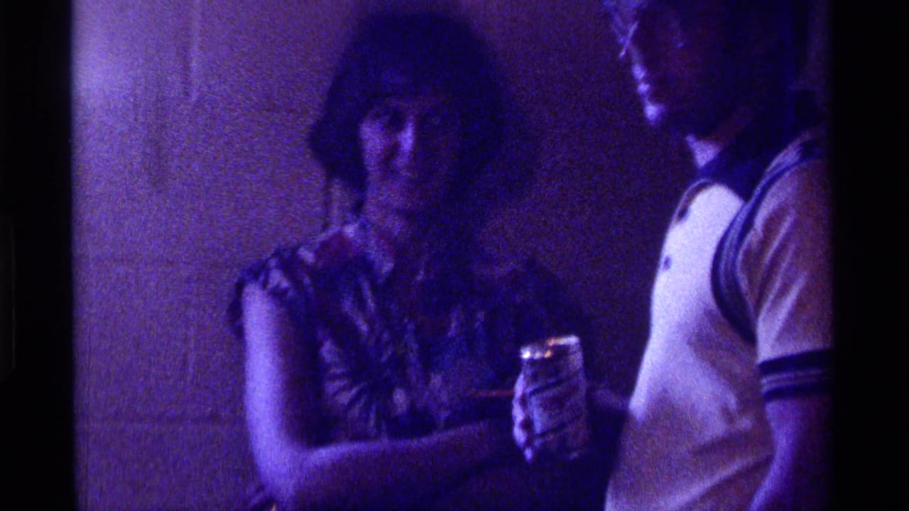 Woman and man standing indoors with beverages under purple-tinted low light.
