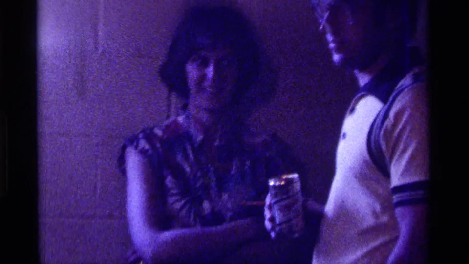 1976 Kentucky home-movie frame of a woman and man at an indoor party, holding drinks in dim purple-tinted light.