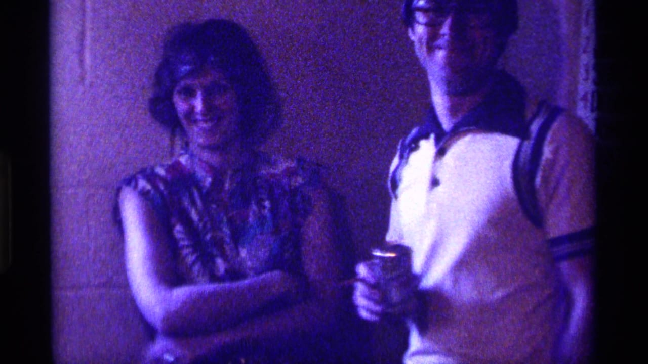 1976 Kentucky home-movie frame showing two young adults smiling indoors with drinks during a casual party.