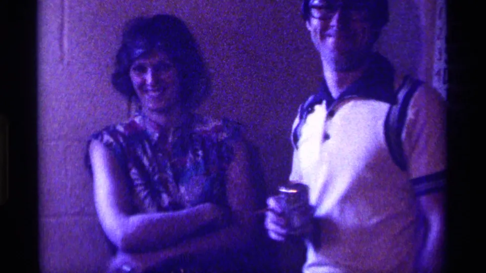 1976 Kentucky home-movie frame showing two young adults smiling indoors with drinks during a casual party.