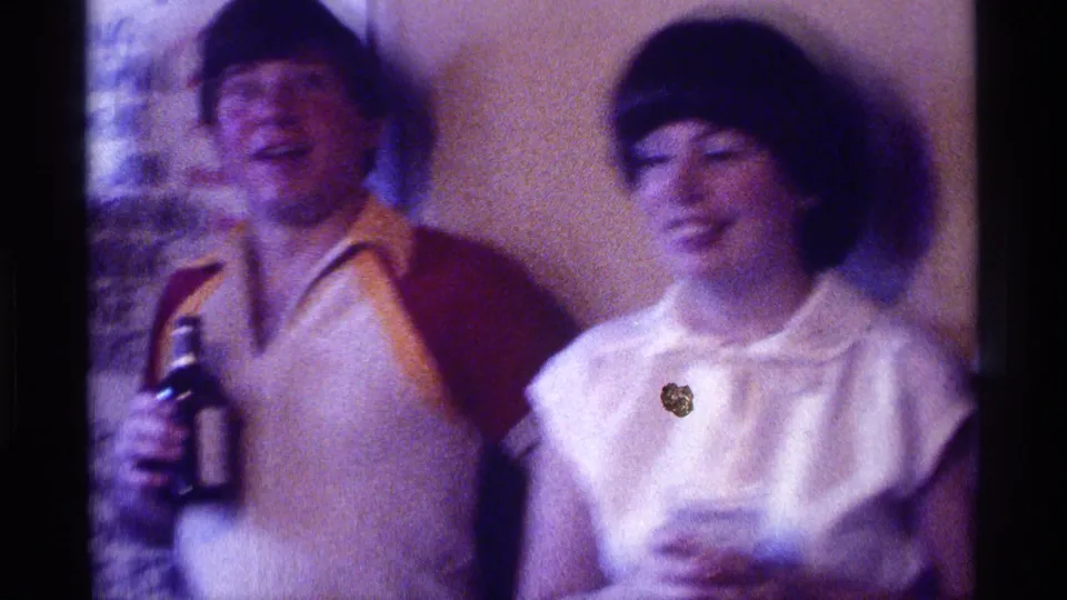 1976 Kentucky home-movie frame of two adults laughing at an indoor party, with drinks and motion blur visible.
