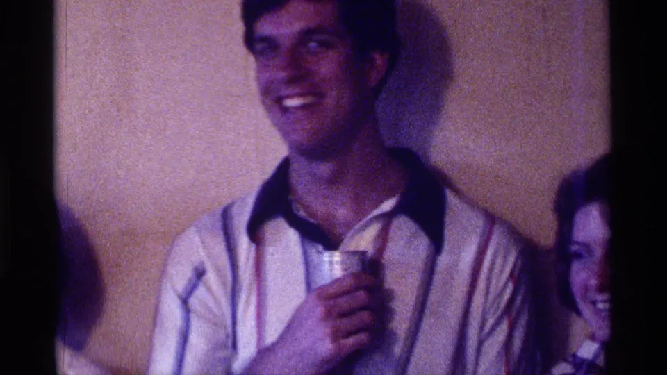 1976 Kentucky home-movie frame of a smiling man with a cup at an indoor party as another face enters the edge.