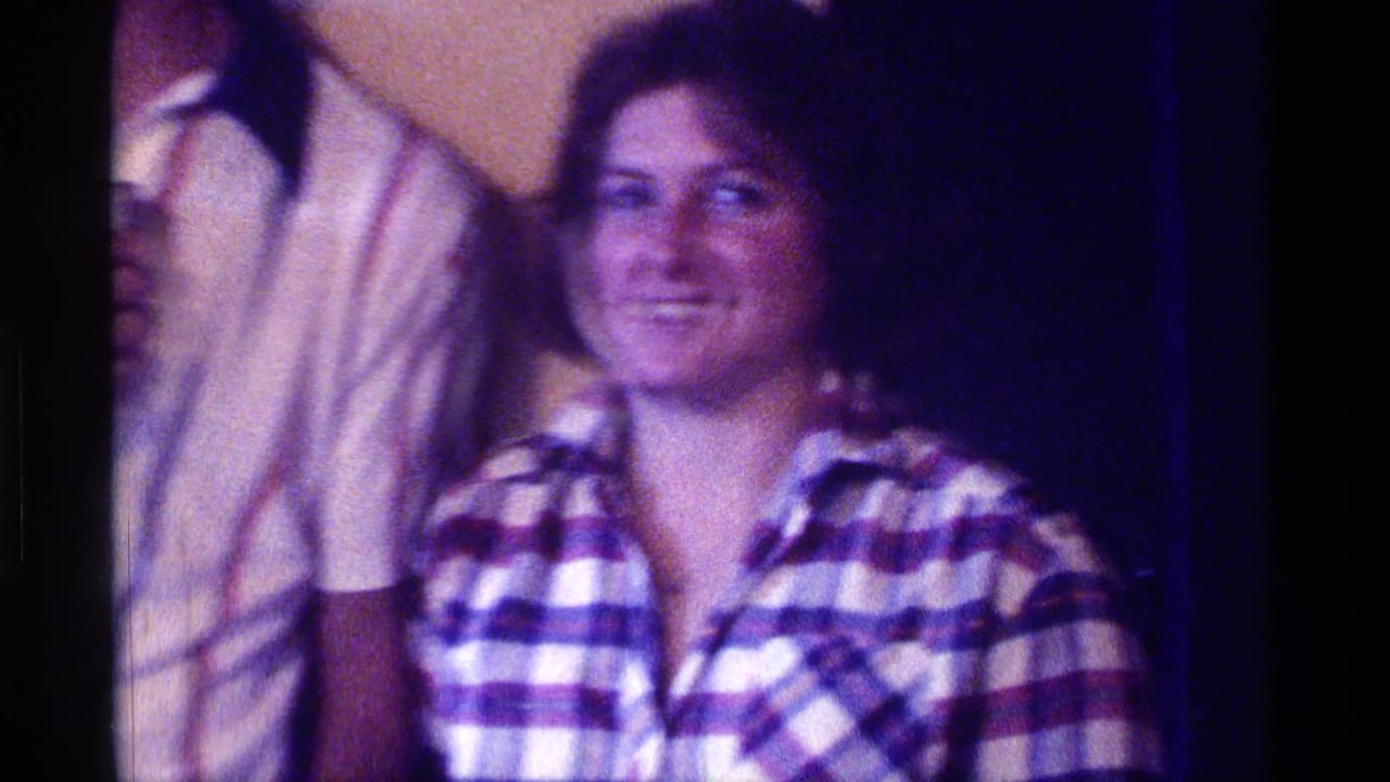 Smiling young woman in a plaid shirt beside partially visible partygoers indoors.