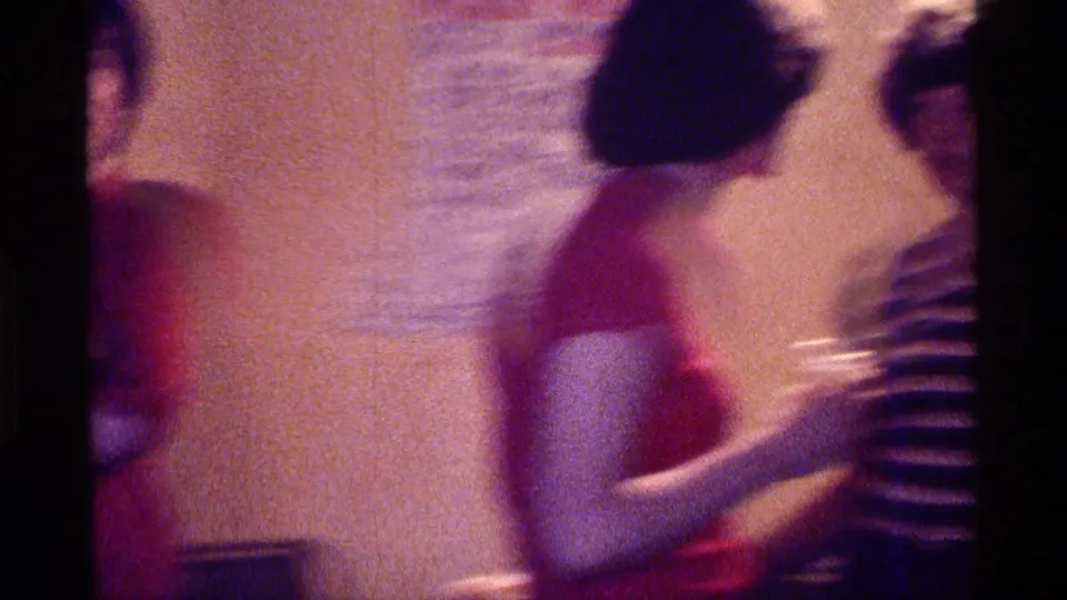 Kentucky 1976 home-movie frame of adults in red and striped shirts talking in a dim private interior.