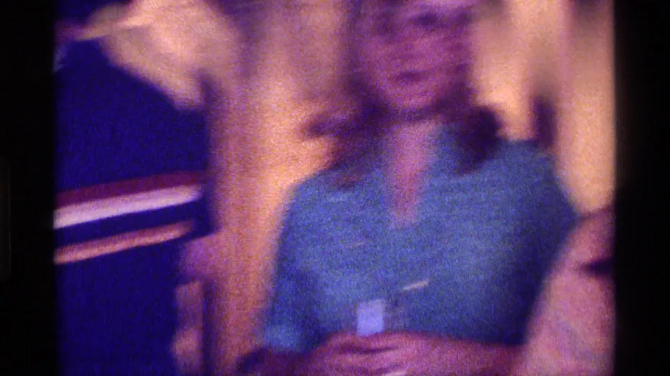 Kentucky 1976 home-movie frame of a young woman in a teal collared shirt holding a drink at an indoor party.