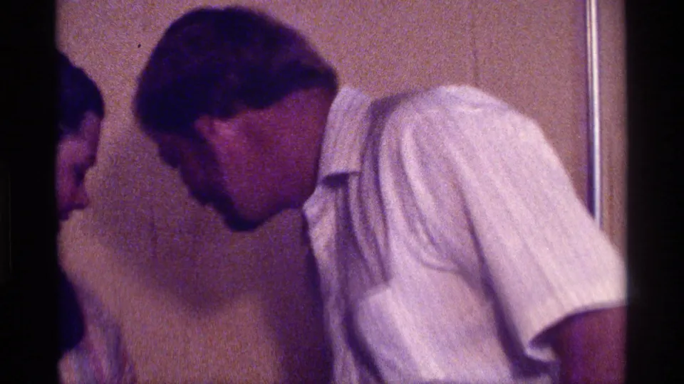 1976 Kentucky home-movie frame of a man in a white shirt leaning toward another adult in a private interior.