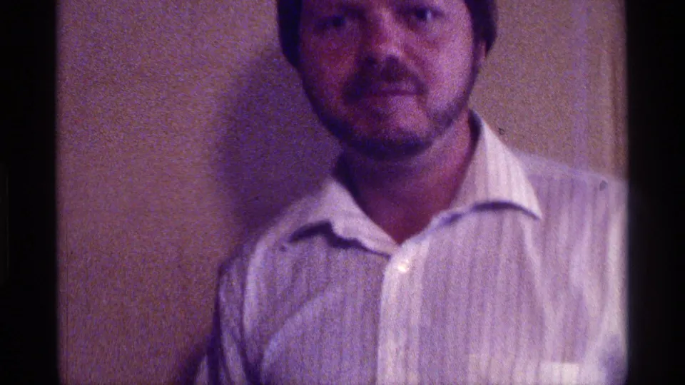 A man in a white collared shirt looks toward the camera in a soft, purple-tinged 1976 Kentucky home-movie frame.