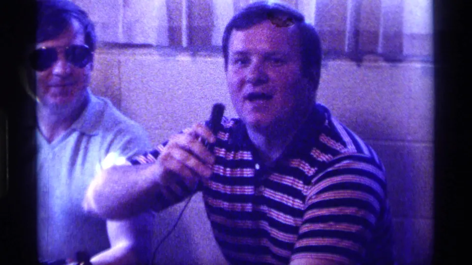 A man in a striped polo speaks into a small microphone beside another man in sunglasses at a 1976 Kentucky indoor party.