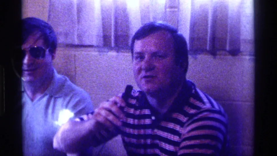 Two adult men sit in a private interior, one holding a small microphone, in grainy 1976 Kentucky 8mm party footage.