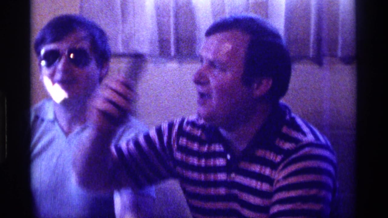 Two adult men indoors, one wearing sunglasses and one in a striped polo holding a microphone.