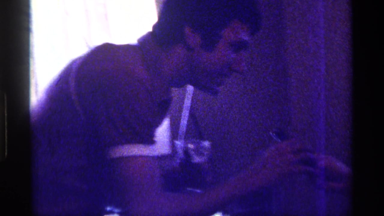 Grainy 1976 Kentucky home-movie frame showing an adult man leaning across a dim private interior near a drink glass with a straw.
