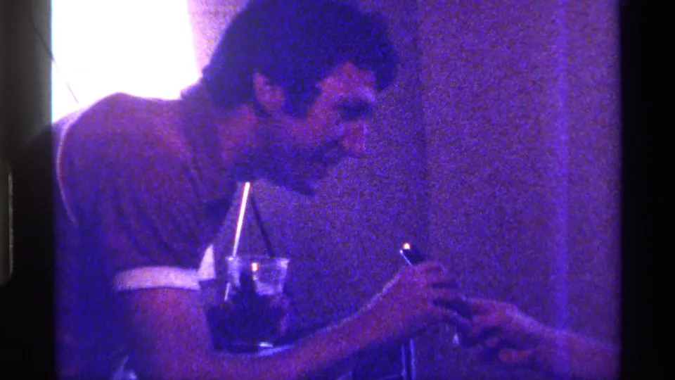 1976 Kentucky home-movie frame of an adult man beside a glass with a straw, appearing to use a lighter near another person's hand.