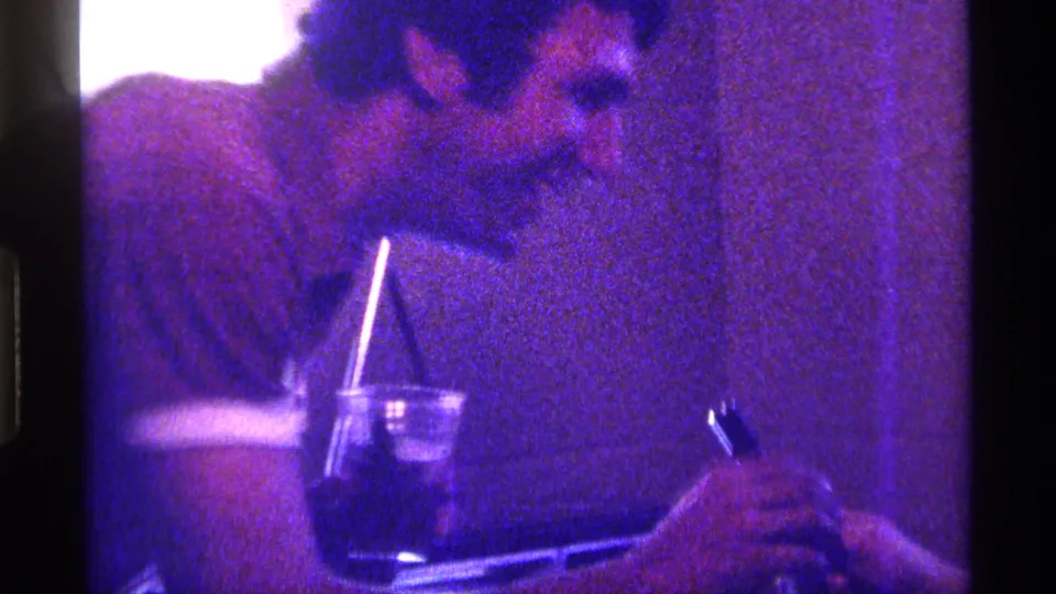 Close 1976 Kentucky home-movie frame of an adult man in a private interior holding a lighter near another hand, with a drink and straw below.