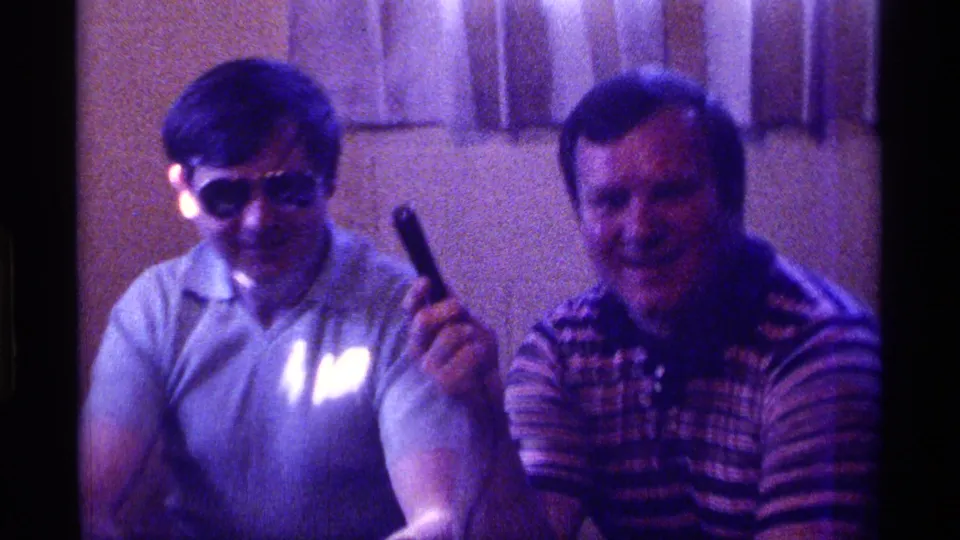 1976 Kentucky home-movie frame of two seated adult men indoors, one wearing sunglasses and holding a microphone while the other smiles.