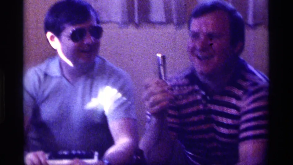 1976 Kentucky 8mm frame showing two adult men at an indoor gathering, with one holding a drink and the other wearing sunglasses.