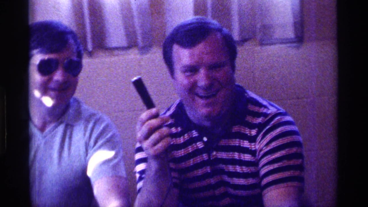 Two adult men seated indoors, one wearing sunglasses and one in a striped shirt holding a black object.