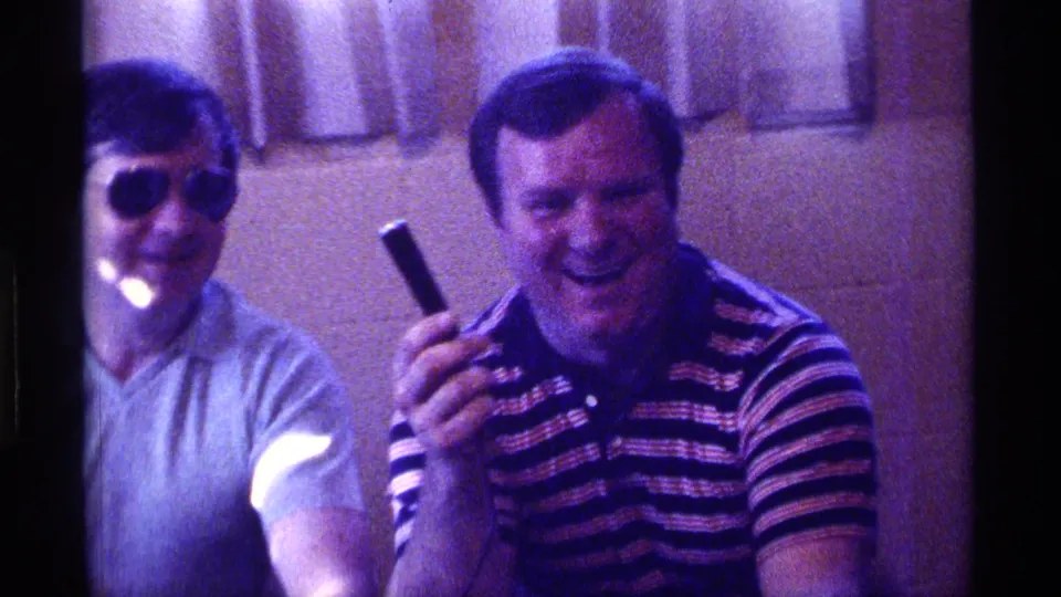 1976 Kentucky home-movie frame of two adult men seated indoors, with one in sunglasses and another smiling in a striped polo while holding a black object.
