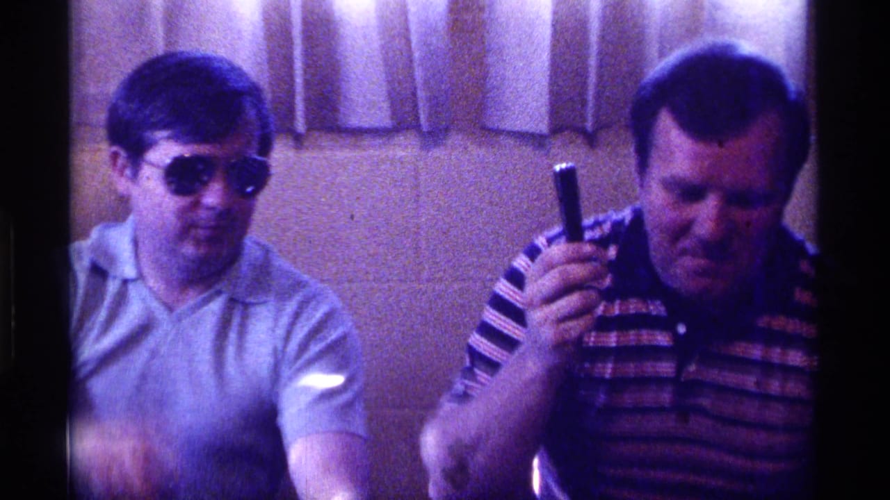 1976 Kentucky indoor frame showing a man in sunglasses beside a man in a striped polo raising a black handheld object near curtains.
