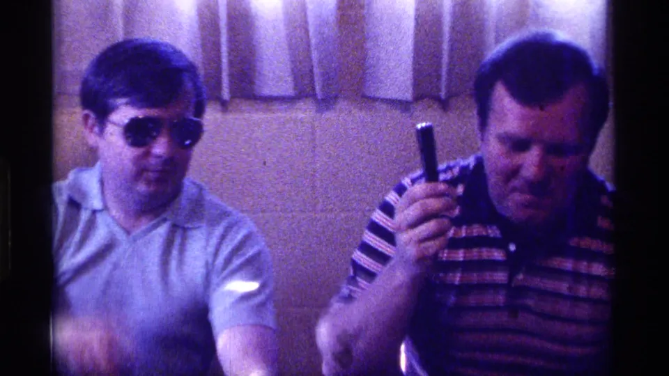 1976 Kentucky indoor frame showing a man in sunglasses beside a man in a striped polo raising a black handheld object near curtains.