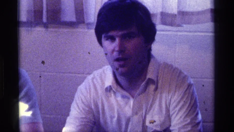 1976 Kentucky home-movie frame of an adult man in a light shirt seated against a block wall and curtains in a dim room.