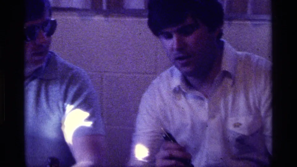 1976 Kentucky indoor frame showing a seated man in a light shirt, with another man in sunglasses partly visible at the edge of the frame.