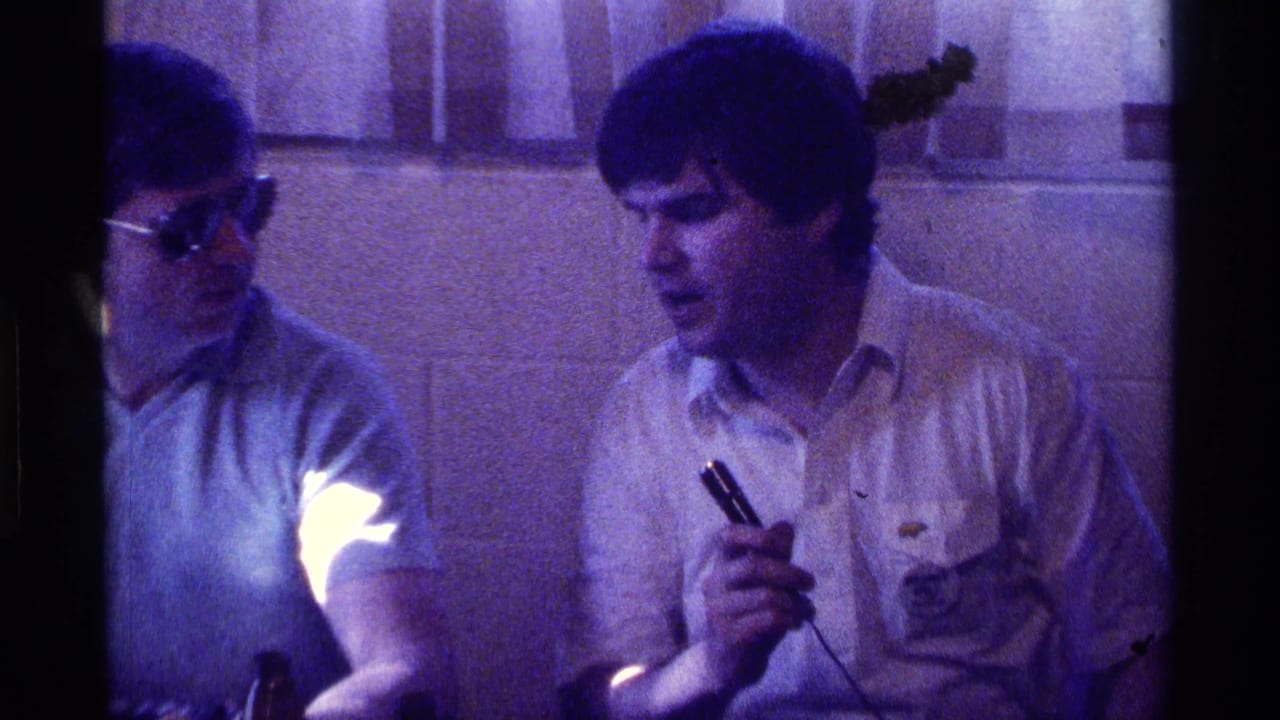 Two adult men sit indoors as one holds a wired microphone near open bottles on a table.