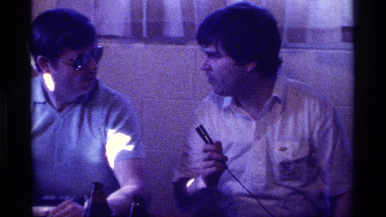 Man in sunglasses faces a man holding a microphone in a dim room with bottles below.