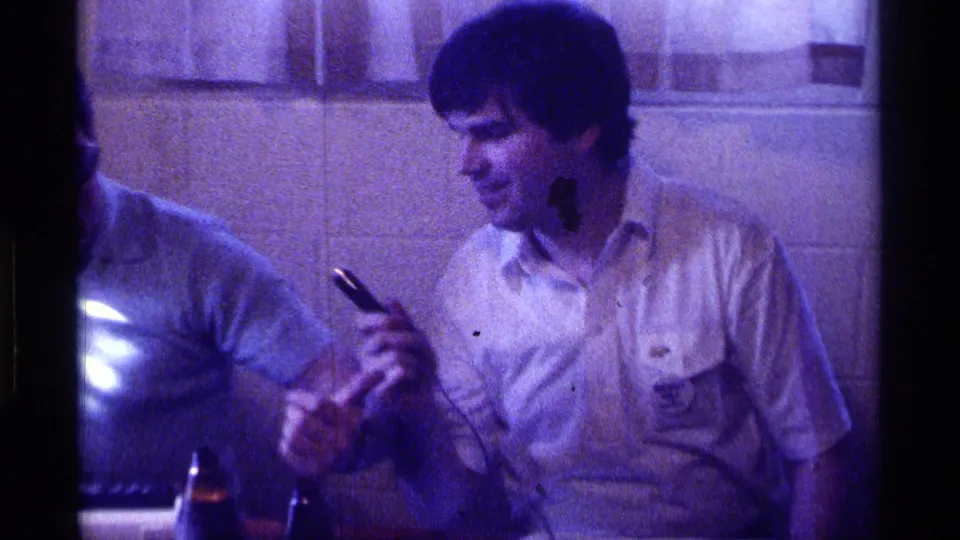 A seated man in a light shirt holds a wired microphone as another adult is partly cropped at left, with bottles visible on the table.