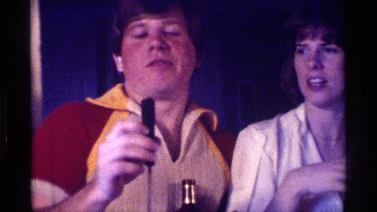 Man holding a microphone stands beside a woman in a dim party interior.
