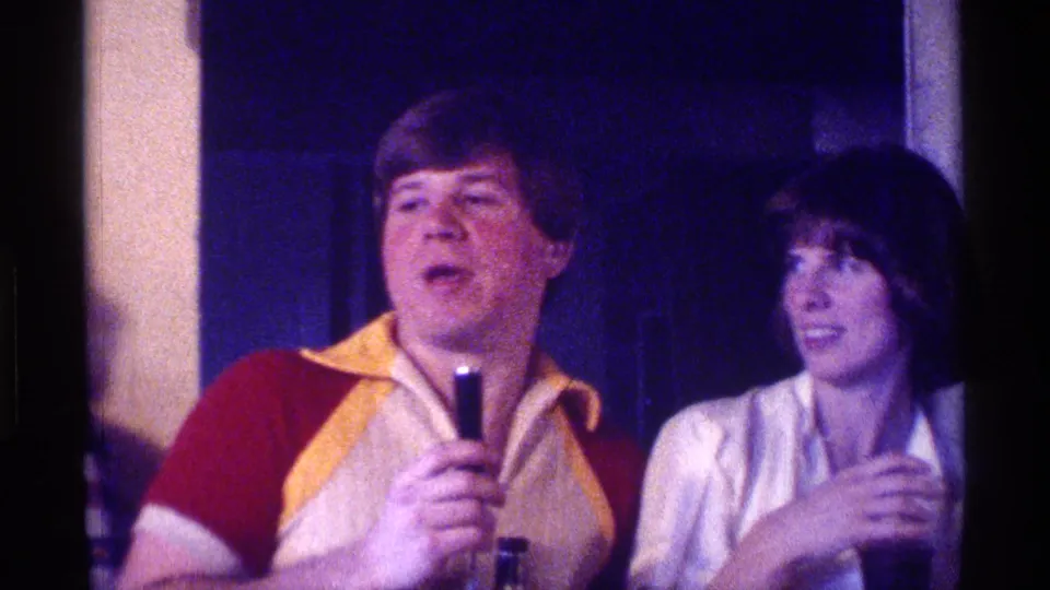 A man holding a microphone stands near a woman who turns toward him, captured in grainy 1976 Kentucky 8mm party footage.