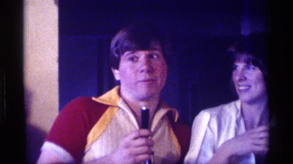 The man holds the microphone at chest height while the woman beside him smiles in the dim 1976 Kentucky home-movie party scene.