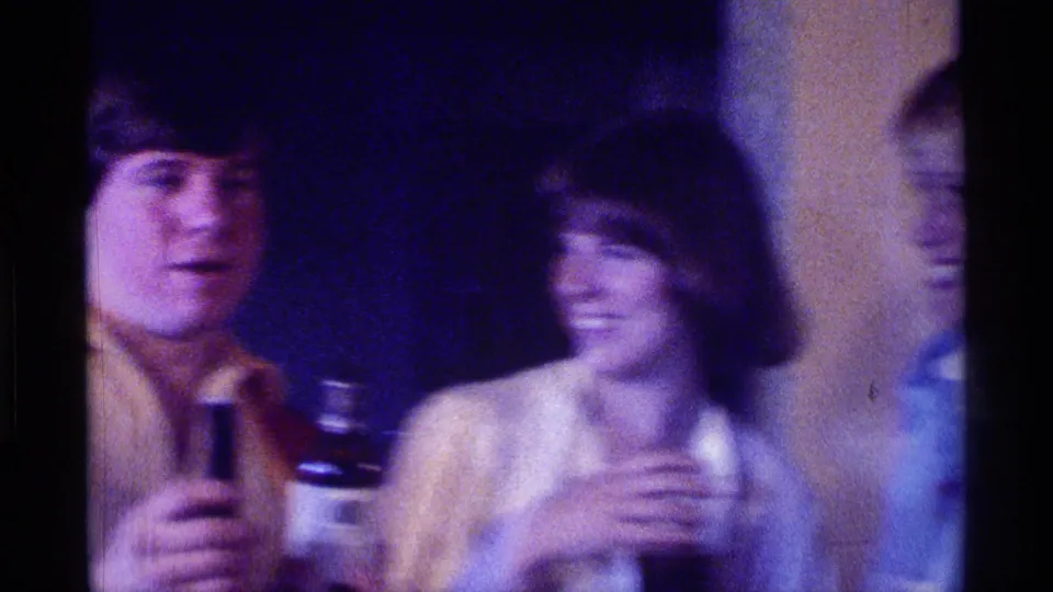 A young man in a collared shirt uses a microphone near a smiling woman in a crowded Kentucky party interior, 1976.