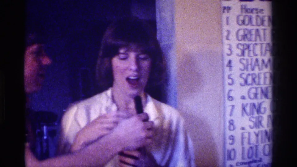 A woman in a light blouse laughs or speaks near a microphone, with a handwritten numbered wall chart visible in the party room.