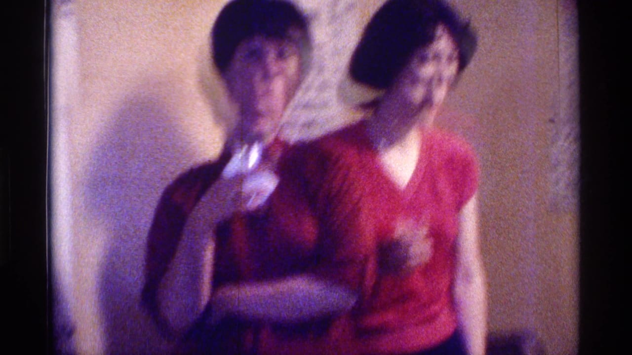 Two women in red tops stand indoors, one holding a drink, with soft 8mm blur.