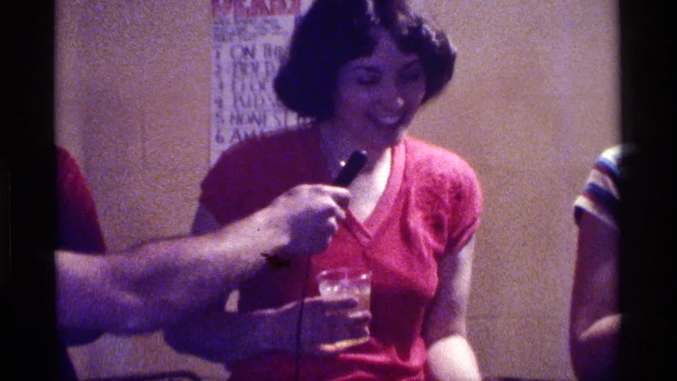 At an indoor Kentucky gathering, a woman in a red top looks down while holding a glass near a handheld microphone.