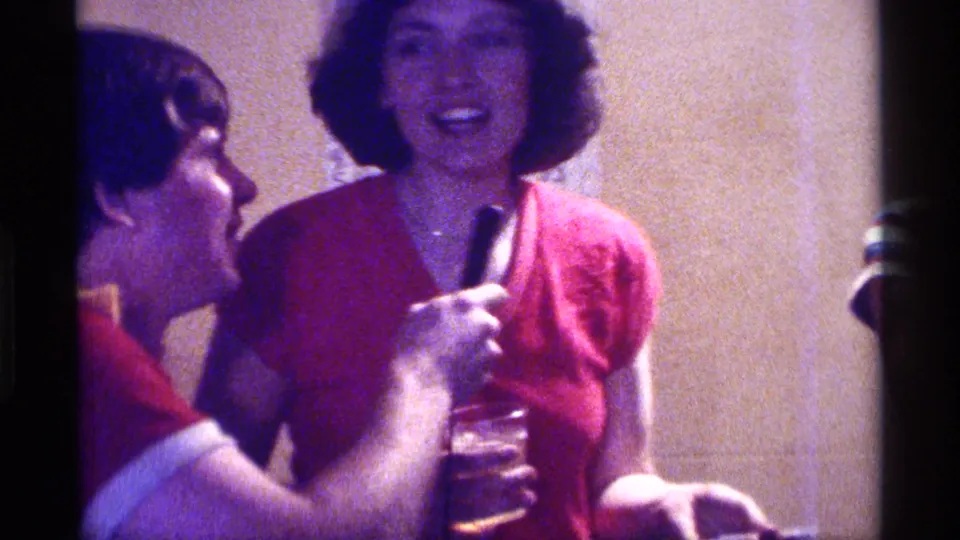 Two women smile and converse near a handheld microphone in 1976 Kentucky home-movie footage of an indoor social gathering.