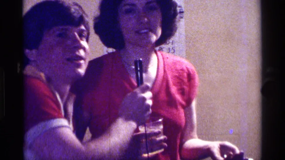 In Kentucky 1976 home-movie footage, a man in a red shirt holds a microphone near a woman in a red top during an indoor gathering.