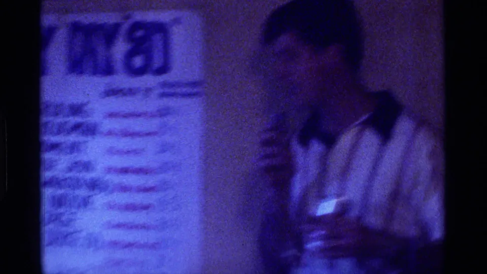 Soft 8mm frame of a man in a white collared shirt standing near a handwritten racing chart in Kentucky, 1976.