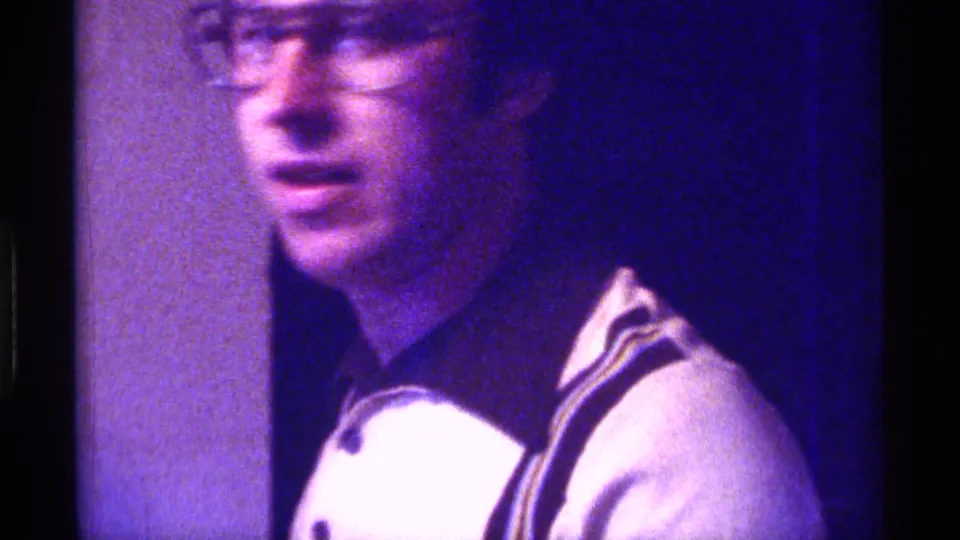 1976 Kentucky 8mm frame of a man with glasses and a white shirt facing the camera in a dim interior.