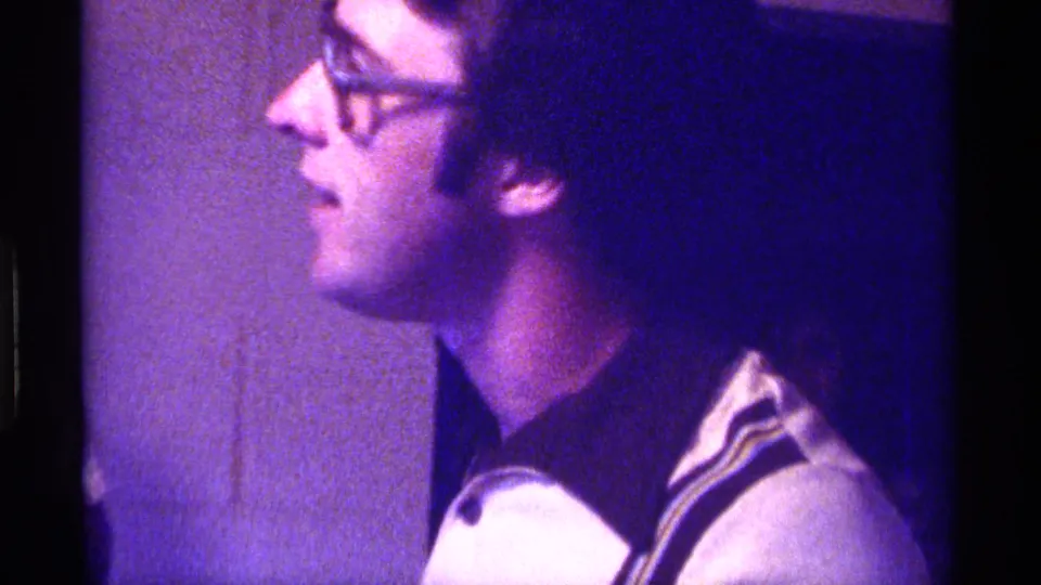 Young man in glasses appears in profile inside a Kentucky home-movie scene from 1976, lit by purple color shift.
