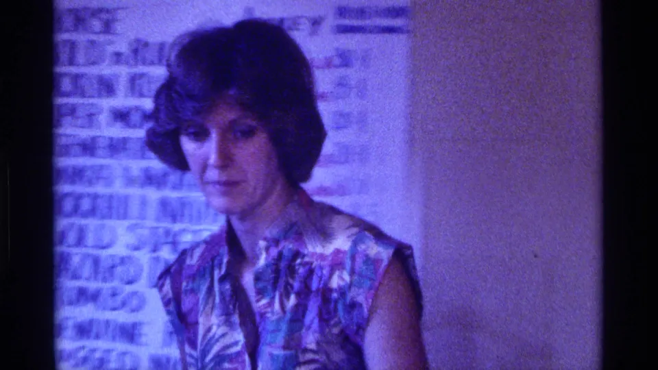 Woman in a patterned sleeveless top stands by a handwritten wall list in a 1976 Kentucky home-movie interior.