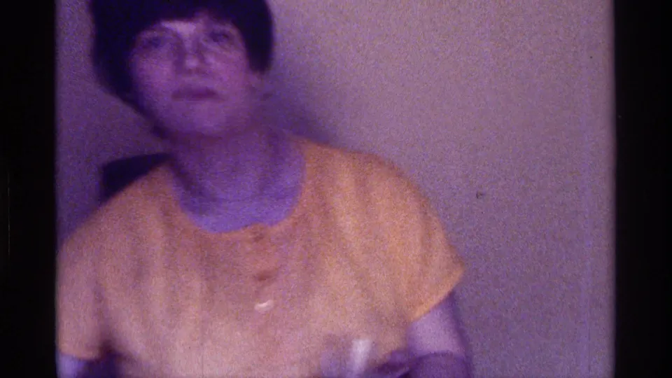 An adult in a yellow shirt sits in a dim Kentucky interior, filmed in grainy 1970s 8mm style with a soft, blurred face.