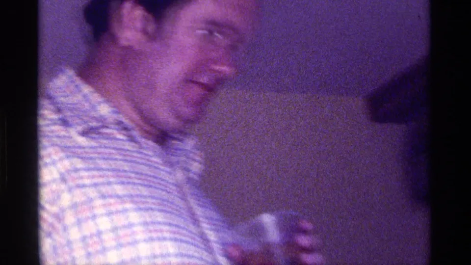 1976 Kentucky 8mm frame showing a smiling man in a striped plaid shirt holding a small glass indoors.