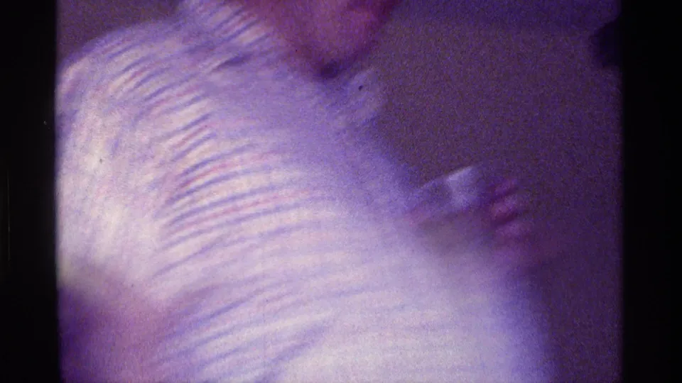 Motion-softened 1976 Kentucky 8mm frame showing a plaid-shirted person with a glass in a dim interior.
