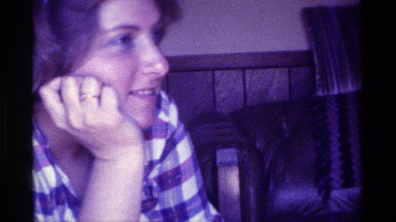 In 1976 Kentucky home-movie footage, a woman in a striped plaid shirt sits close to the camera with her chin on her hand in a softly lit private interior.