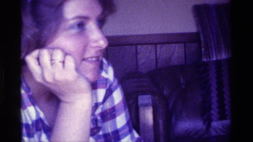 In 1976 Kentucky home-movie footage, a woman in a striped plaid shirt sits close to the camera with her chin on her hand in a softly lit private interior.