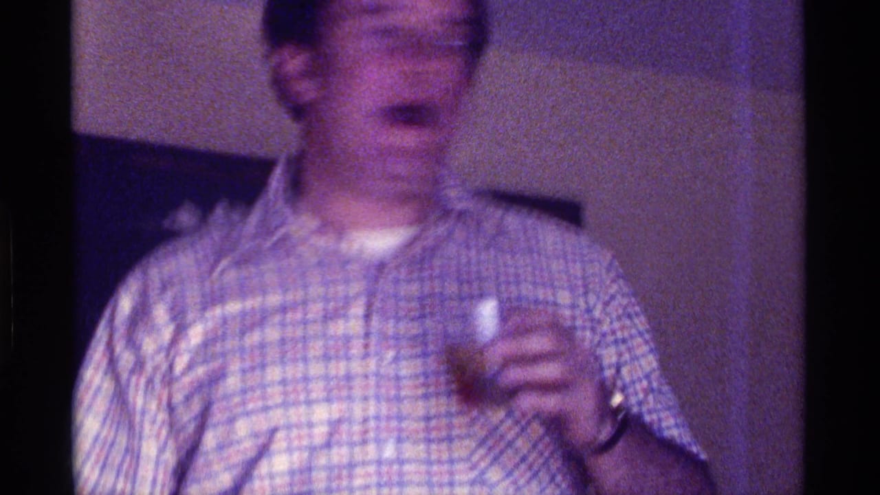 A man in a plaid shirt holds a small drink in a dim Kentucky home interior, filmed in soft-focus 8mm footage from 1976.